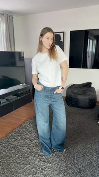 Valkoinen t-paita yhdistettynä leveälahkeisiin sinisiin farkkuihin, joissa on klassinen malli ja kevyt haalistunut efekti. Taustalla on musta sohva ja televisio.