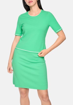 Frau trägt ein grünes Kleid mit kurzen Ärmeln und weißem Besatz an Ärmeln und Taille, posiert mit einer Hand auf der Hüfte vor weißem Hintergrund.