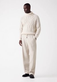 Gruby kremowy sweter z golfem o fakturze z wzorem w diamenty, zestawiony z jasnobeżowymi szerokimi spodniami i czarnymi butami.
