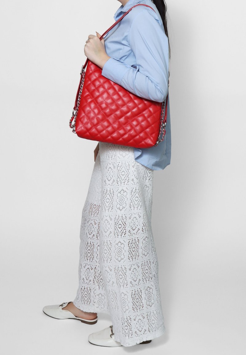 Borsa a tracolla in pelle trapuntata rossa con catene argentate, forma quadrata grande. Indossata con una camicia blu e pantaloni con motivo in pizzo bianco.