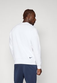 Sweatshirt blanc Nike à manches longues, avec des poignets et un ourlet côtelés. Logo discret bleu marine à l'arrière près de l'ourlet. Texture lisse et coupe décontractée.