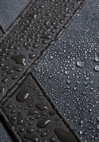Tessuto nero resistente all'acqua con gocce, caratterizzato da una superficie testurizzata e spallacci lisci. Luce notevole sulle gocce d'acqua.