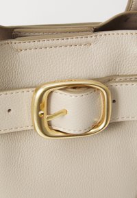 Détail d'un sac à main en cuir beige mettant en avant une boucle en métal doré, une texture lisse et des coutures visibles. La boucle est de forme carrée avec une goupille.