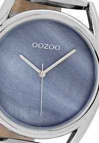 Montre OOZOO ronde avec un cadran dégradé bleu, aiguilles argentées et logo. Boîtier en métal avec une finition polie. Design simple et élégant.