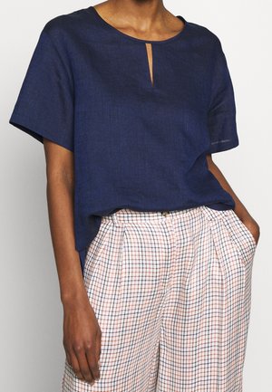 Persoon draagt een marineblauw T-shirt met korte mouwen, een klein sleutelgat aan de voorkant, en witte geruite high-waist broek met handen in de zakken.