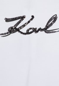 Camiseta de tela blanca con un diseño en negro de letras de script con lentejuelas que dice "Karl", con una textura brillante y letras curvadas.