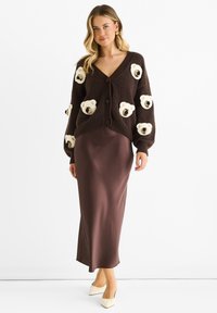 Brown knit cardigan with bear appliqués, v-neck, and oversized sleeves, paired with a brown satin midi skirt and beige heel shoes.