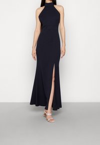 Vestido maxi azul marino con escote halter, abertura frontal, cuerpo ajustado y falda fluida; combinado con tacones plateados de tiras. Textura de tela suave.