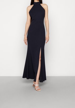 Vestido maxi azul marino con escote halter, abertura frontal, cuerpo ajustado y falda fluida; combinado con tacones plateados de tiras. Textura de tela suave.