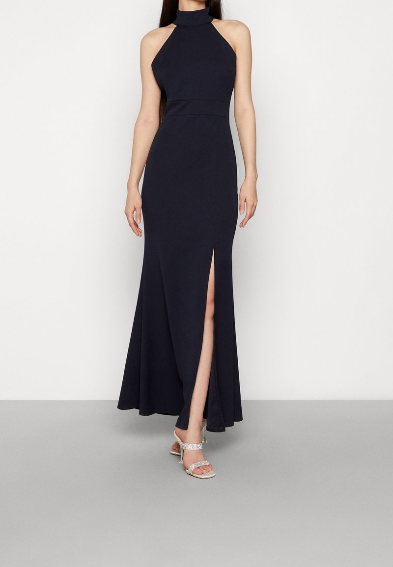 Vestido maxi azul marino con escote halter, abertura frontal, cuerpo ajustado y falda fluida; combinado con tacones plateados de tiras. Textura de tela suave.