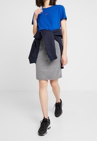 Modrý Nike tričko s logem, šedá kostkovaná sukně, černé sportovní boty. Tmavý svetr přehozený přes ramena. Hladká tkanina a přiléhavý střih.