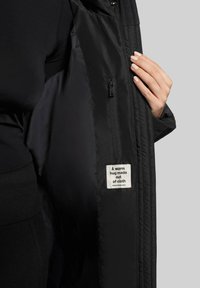 Die Innenseite der schwarzen Jacke verfügt über eine Reißverschlusstasche und ein Etikett mit der Aufschrift: "Eine warme Umarmung aus Stoff." Glatte Textur, strapazierfähiges Material.