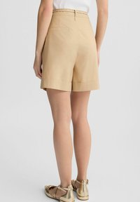 Bermuda beige con una vestibilità su misura, orlo risvoltato e passanti per cintura intrecciati. Tessuto liscio con due tasche posteriori. Indossate con sandali dorati.