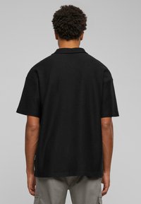 Black polo shirt with short sleeves, textured fabric, classic collar, and loose fit. Visible tag on the side, worn with light gray cargo pants.