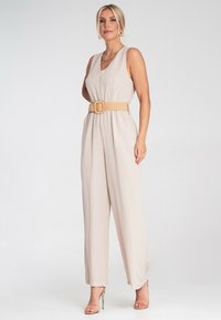 Beżowy kombinezon bez rękawów, z wcięciem w talii ozdobionym plecionym paskiem oraz szerokimi nogawkami. Gładki, lekki materiał z subtelnym opadem.