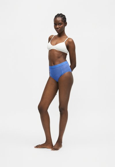 Γυναίκα στέκεται ξυπόλυτη, φοράει λευκό μπραλέτα και μπλε εσώρουχα με ψηλή μέση σε λευκό φόντο.