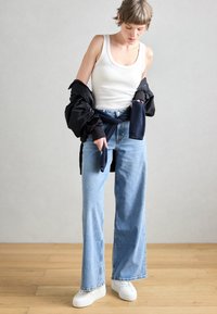 Λευκό ριγέ αμάνικο τοπ, μαύρο μπουφάν puffer με σούρες στα μανίκια, ανοιχτό μπλε τζιν παντελόνι φαρδύ και λευκά αθλητικά παπούτσια, στέκεται σε ξύλινο πάτωμα.