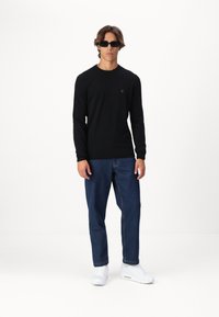 Schwarzes Langarmshirt mit rundem Halsausschnitt, kombiniert mit blauen Jeans und weißen Sneakers. Modell trägt schwarze Sonnenbrillen.