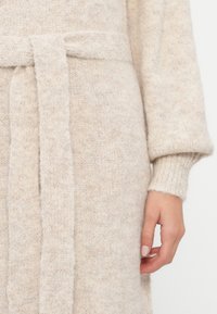 Cardigan beige lavorato a maglia con finitura testurizzata, dotato di una cintura in tessuto in vita e polsini a costine.