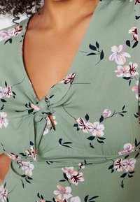 Nahaufnahme eines grünen Blumenkleides mit pinken und weißen Blumen, das ein vorn gebundenes Detail und kurze Ärmel aufweist, getragen von einer Person.