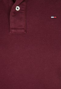 Burgundin värinen puuvillapolo-paita, jossa on teksturoitu pinta, kahden napin nappilista ja pieni brodeerattu logo punaisella, valkoisella ja sinisellä.