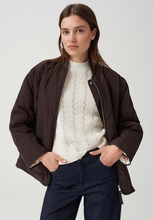 Giovane donna che indossa una giacca trapuntata marrone scuro sopra un maglione bianco lavorato a maglia e jeans scuri, in piedi con le mani in tasca contro uno sfondo chiaro.