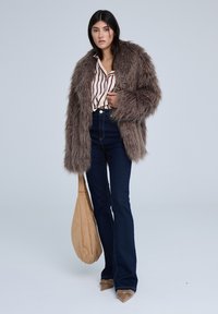 Cappotto in pelliccia sintetica color marrone chiaro, camicia a righe bianca e rossa, jeans scuri a vita alta, borsa beige e tacchi nude a punta; texture liscia.