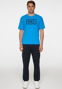 Sininen puuvillapaita lyhyillä hihoilla ja musta "SKI" -grafiikka edessä, yhdistetty tummiin housuihin ja valkoisiin lenkkikengiin.