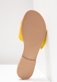 Semelle d'une sandale jaune à bout ouvert avec une semelle en caoutchouc beige texturée, photographiée debout contre un fond blanc en angle.