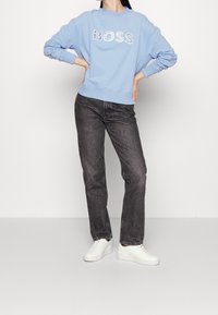 Ljusblå sweatshirt med "BOSS"-logotyp i vitt, matchad med mörkgrå jeans. Modellen har vita sneakers och står med händerna på höfterna.