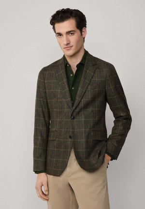 Brauner karierten Blazer aus strukturiertem Stoff, mit Reverskragen, zwei Fronttaschen und einem einzelnen Knopfverschluss. Kombiniert mit einem dunkelgrünen Hemd.