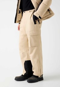 Beige Cargo-Hosen mit mehreren Taschen, einem entspannten Schnitt und einem schwarzen Einsatz am Knöchel. Das Material wirkt strapazierfähig und glatt.