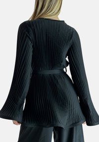 Top negro con plisado, mangas largas y acampanadas, y cinturón atado en la cintura. Tejido suave y brillante con un patrón vertical texturizado.