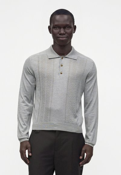 Homme portant un polo en maille gris clair à manches longues avec un devant à motifs et un pantalon foncé, debout devant un fond uni.