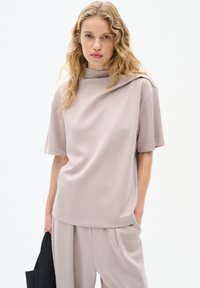Halenka s krátkým rukávem v světle šedohnědé barvě z měkké tkaniny, s imitací stojáčku a volným střihem, zdobená drapovaným detailem na rameni.
