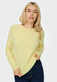 Pull jaune clair avec un col rond côtelé, des manches longues et une texture douce. Tenu légèrement en bas avec les mains dans les poches d'un jean bleu.