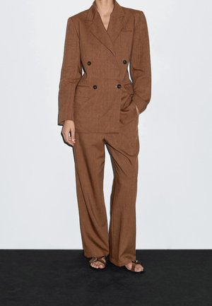 Personne portant un blazer marron croisé et un pantalon large assorti, ainsi que des sandales ouvertes, se tenant devant un mur blanc uni.