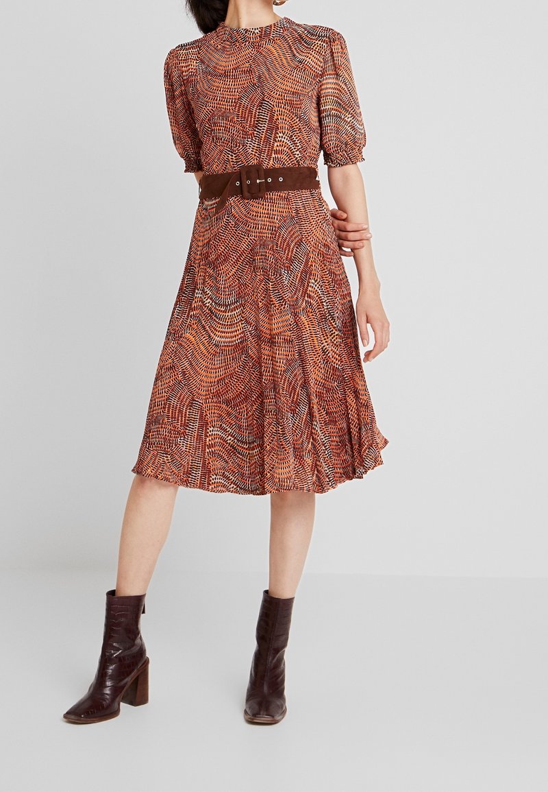 Robe imprimée à manches bouffantes courtes, présentant un motif orange et marron, cintrée par une ceinture marron. Associée à des bottes à talons marron.