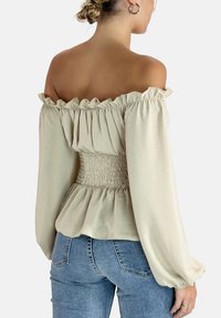 Hellbeige schulterfreies Oberteil mit rüschigem Ausschnitt und langen Ärmeln, ausgestattet mit einem smockierten Taillenbereich und einem schwingenden Saum. Der Stoff hat eine weiche Textur.