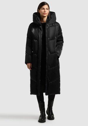 Sort pufferjakke med hætte, der har et langt design, quiltet tekstur, front knaplukning og sidelommer. Båret over et sort outfit.