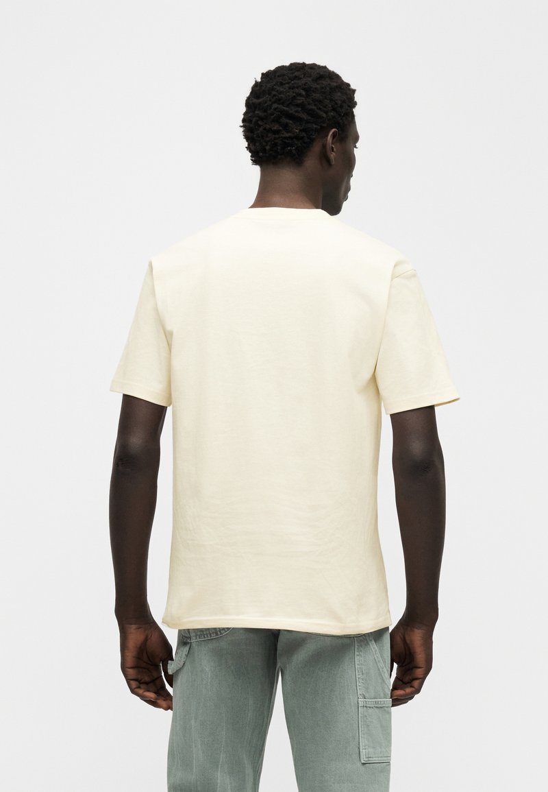 T-shirt di cotone color crema con maniche corte, scollo rotondo e vestibilità rilassata, vista da dietro. Accompagnata da pantaloni verde chiaro.