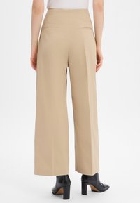 Wide-leg trousers in light beige fabric with a smooth texture, featuring two back pockets and a high waistband. Worn with black heeled boots.
