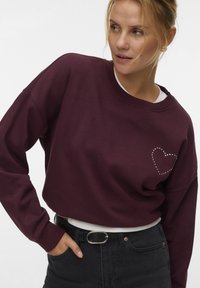 Vrouw in een bordeauxrode sweatshirt met een nagestikte hartontwerp, zwarte jeans en een zwarte riem, die iets opzij kijkt.