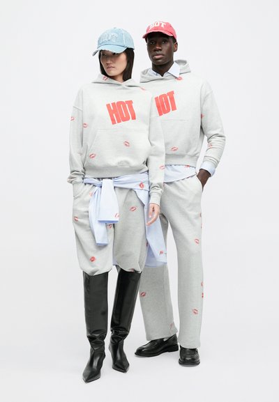 Två modeller bär gråa hoodies och mjukisbyxor med röd text "HOT" och läpptryck, kombinerade med kepsar och svart skor, stående sida vid sida.