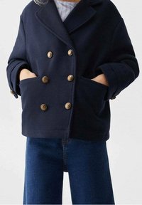 Mörkblå dubbelknäppt jacka med guldknappar, två framfickor och en strukturerad tyg. Åtföljs av jeans i denim.