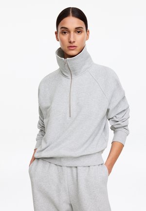 Kvinde iført lysegrå, langærmet sweatshirt med høj krave og lynlås samt matchende joggingbukser med hænderne i lommerne mod hvid baggrund.