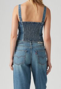 Bakifrån av en person som bär en blå denimsmockad tank top och högmidjade blå jeans med bakfickor.
