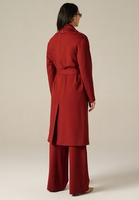 Long manteau rouge avec une ceinture, présentant un col haut, un dos fendu et une texture lisse, associé à un pantalon rouge à jambes larges.