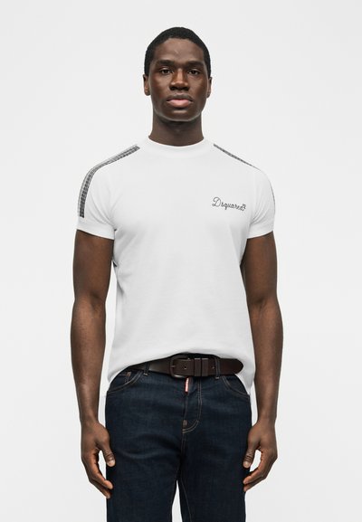 Νεαρός άνδρας που φοράει λευκό T-shirt Dsquared2 με ριγέ λεπτομέρειες στους ώμους και σκούρο μπλε τζιν, στέκεται μπροστά σε απλό φόντο.