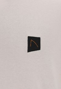 Tissu gris clair avec un petit patch rectangulaire noir présentant un dessin linéaire abstrait orange, cousu au centre.